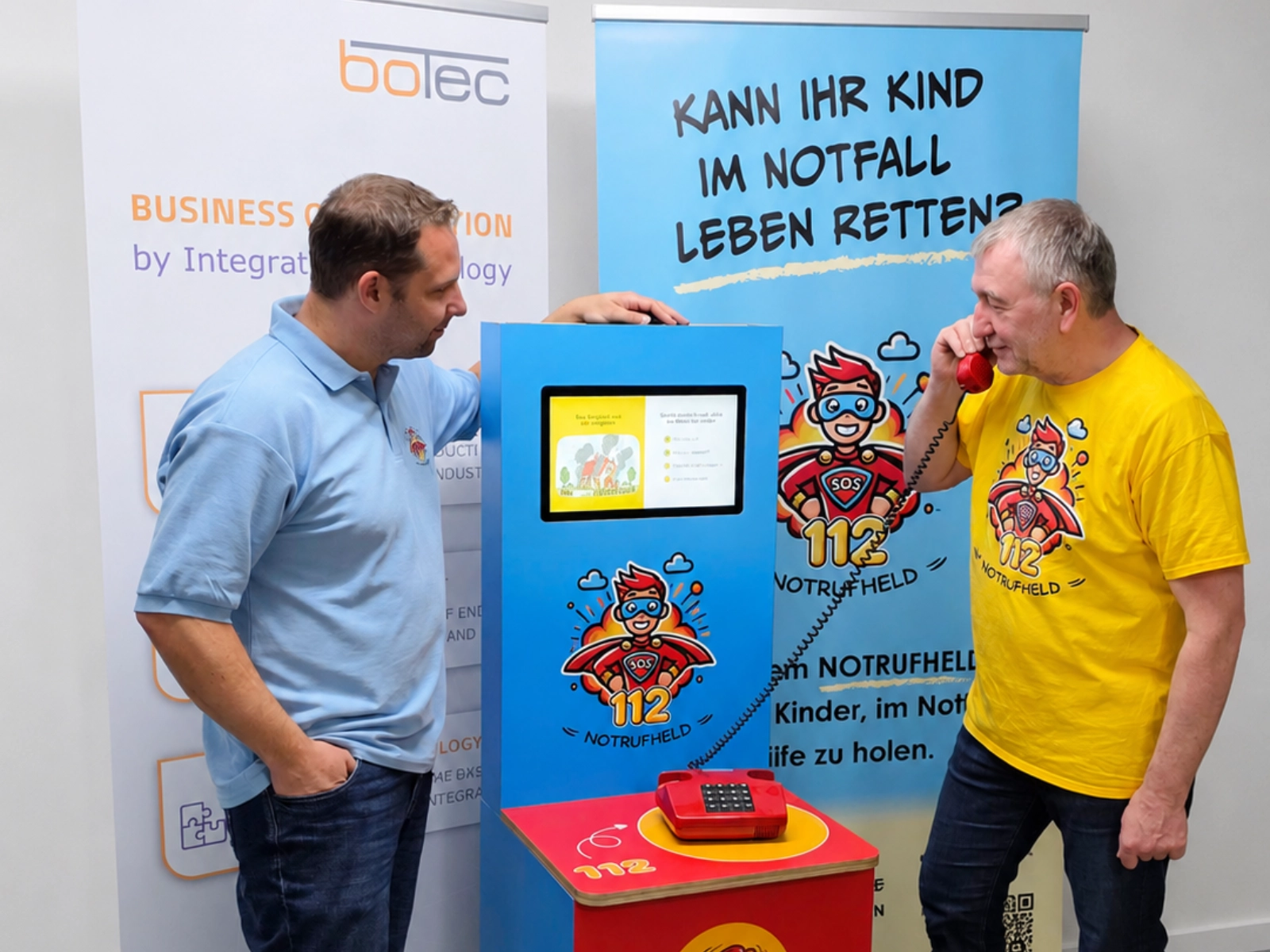 botec-sponsoring Uwe Rupp von botec GmbH und Christian Bleser von blecon stehen neben einem Notrufheld Lernsystem. Uwe telefoniert mit der Leitstelle und testet unser Lernsystem